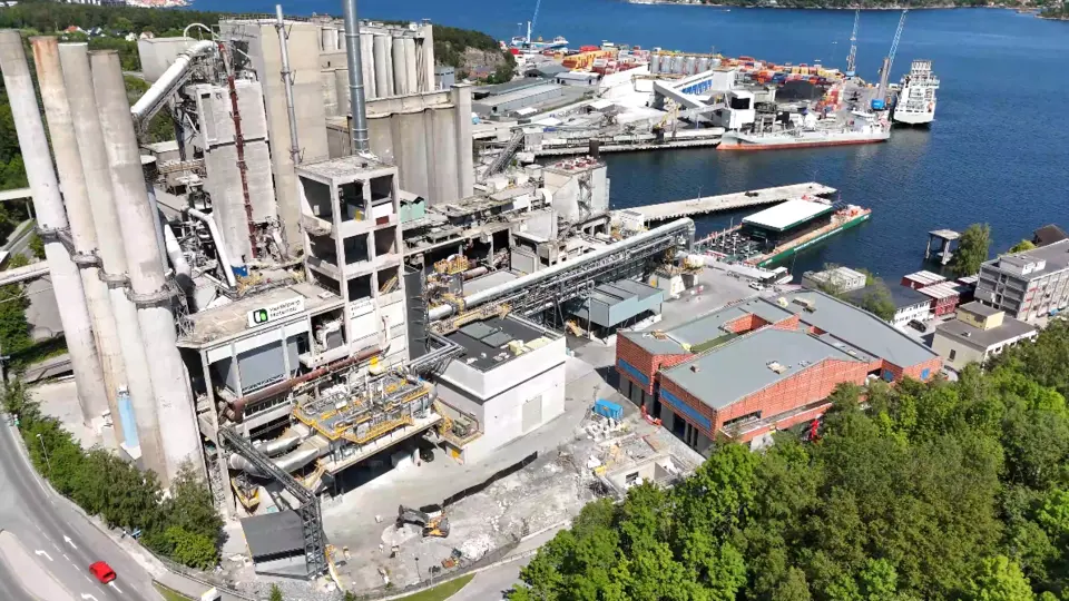 Aerial view of an industrial facility by a waterfront, featuring tall concrete silos on the left, a central processing plant, and warehouses with red roofs to the right. A harbour with docked ships and containers is visible in the background, while trees and a road border the bottom of the image.