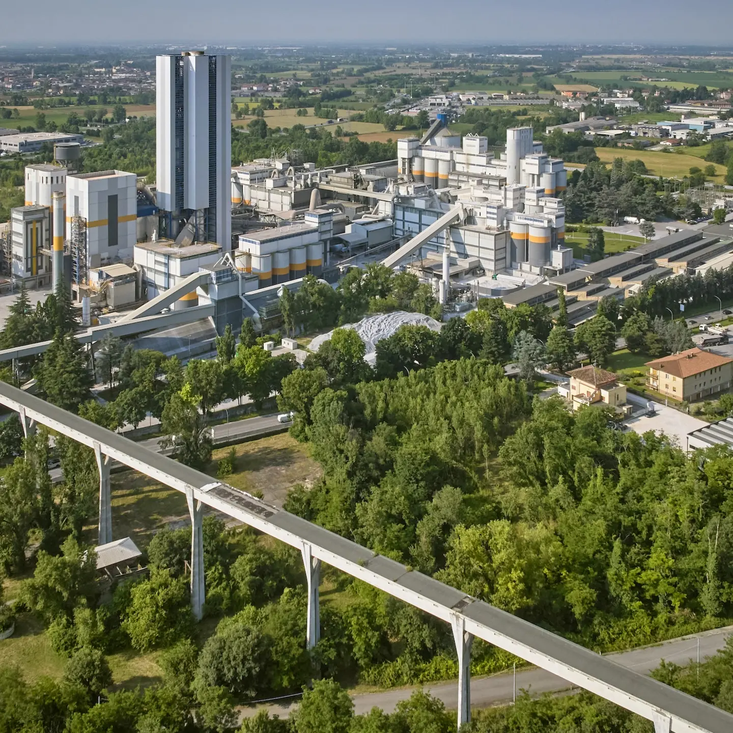 Aerial view of a cement plant surrounded by greenery, with tall silos and factory buildings in the center. A long elevated conveyor system crosses the foreground, connecting different sections of the site. 