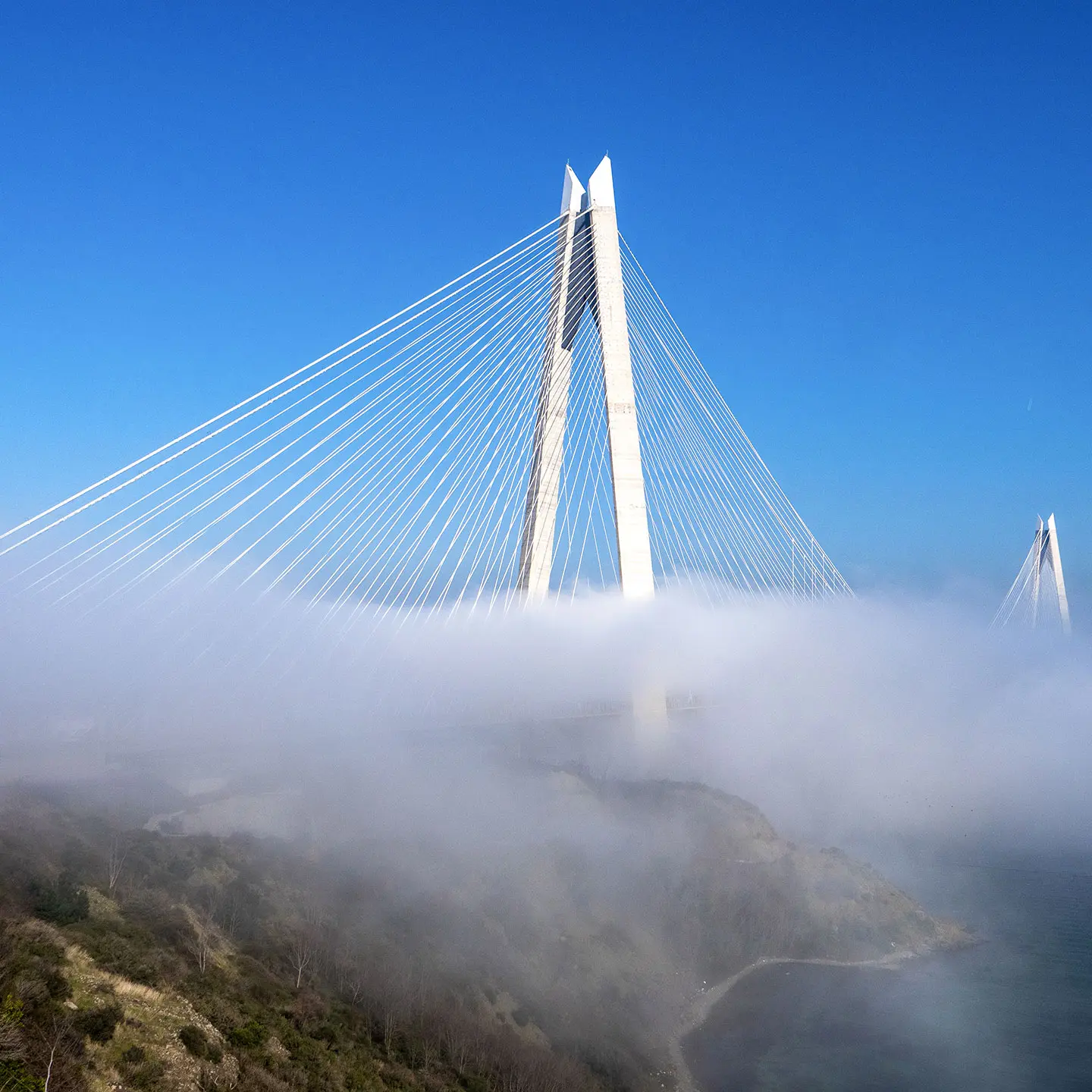 Cable-stayed bridge with tall white pylons partially covered in fog, spanning over a river and connecting cliffs with greenery, under a clear blue sky.