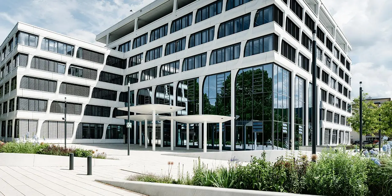 Hauptverwaltung von Heidelberg Materials: Modernes Bürogebäude mit großen Glasfassaden, flankiert von Bäumen auf der linken Seite und Flaggen rechts. Im Vordergrund ein gepflasterter Eingangsbereich mit Grünflächen und niedrigen Lampen.