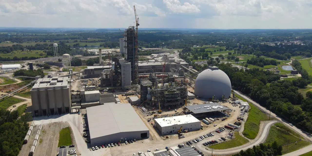 A large cement plant under construction, with several buildings and a silo visible. Cranes and other construction equipment are on site.