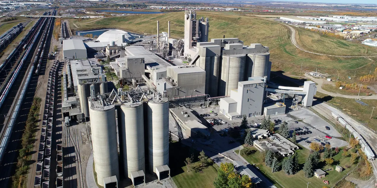 Aerial view of a cement plant with multiple storage silos, towers, and buildings. Train tracks run alongside the facility.