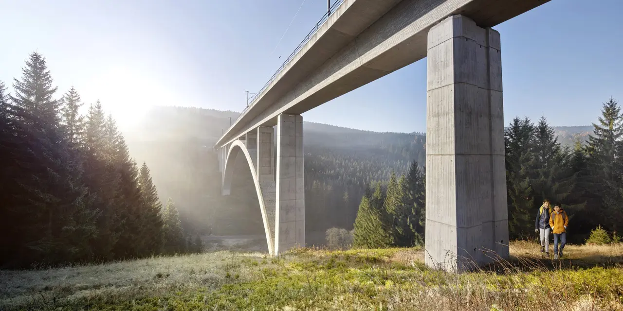 A bright grey bridge in the forest shrouded in fog, two hikers are walking under the bridge