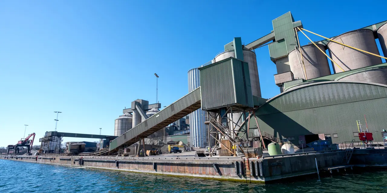 Ein weitläufiger Industriekomplex mit hohen Silos und umfangreichen Förderbändern liegt an einem Gewässer, alles unter der Weite eines klaren blauen Himmels. 