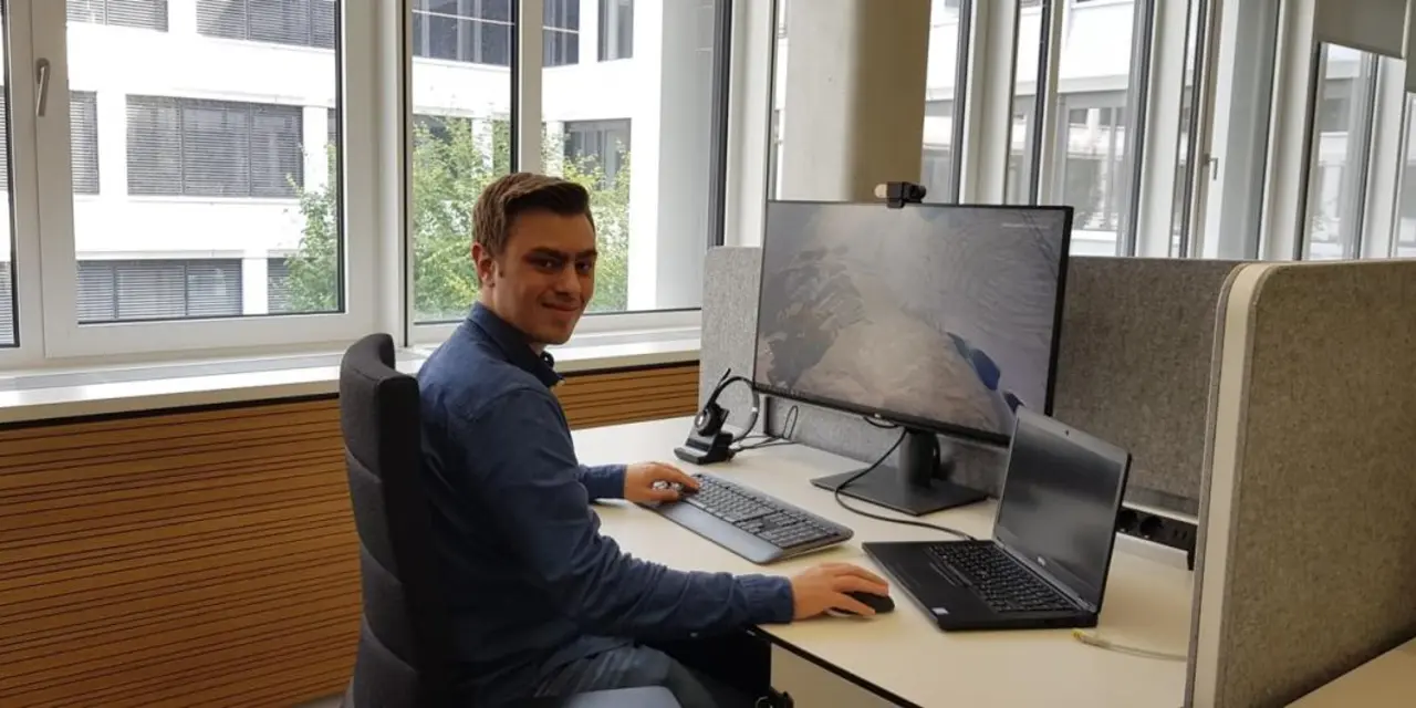 Junger Mann an einem Schreibtisch mit Laptop, Bildschirm, Tastatur und Headset