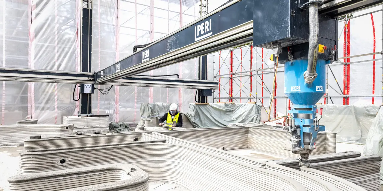 Das Bild zeigt eine große Druckmaschine, die an einem Gerüst befestigt ist und einen teigartigen Beton aus einer Düse presst.