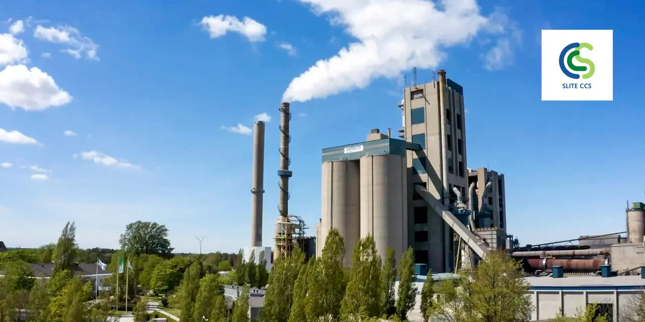 A cement plant with two chimneys, trees in front of it, blue sky with a few clouds behind it