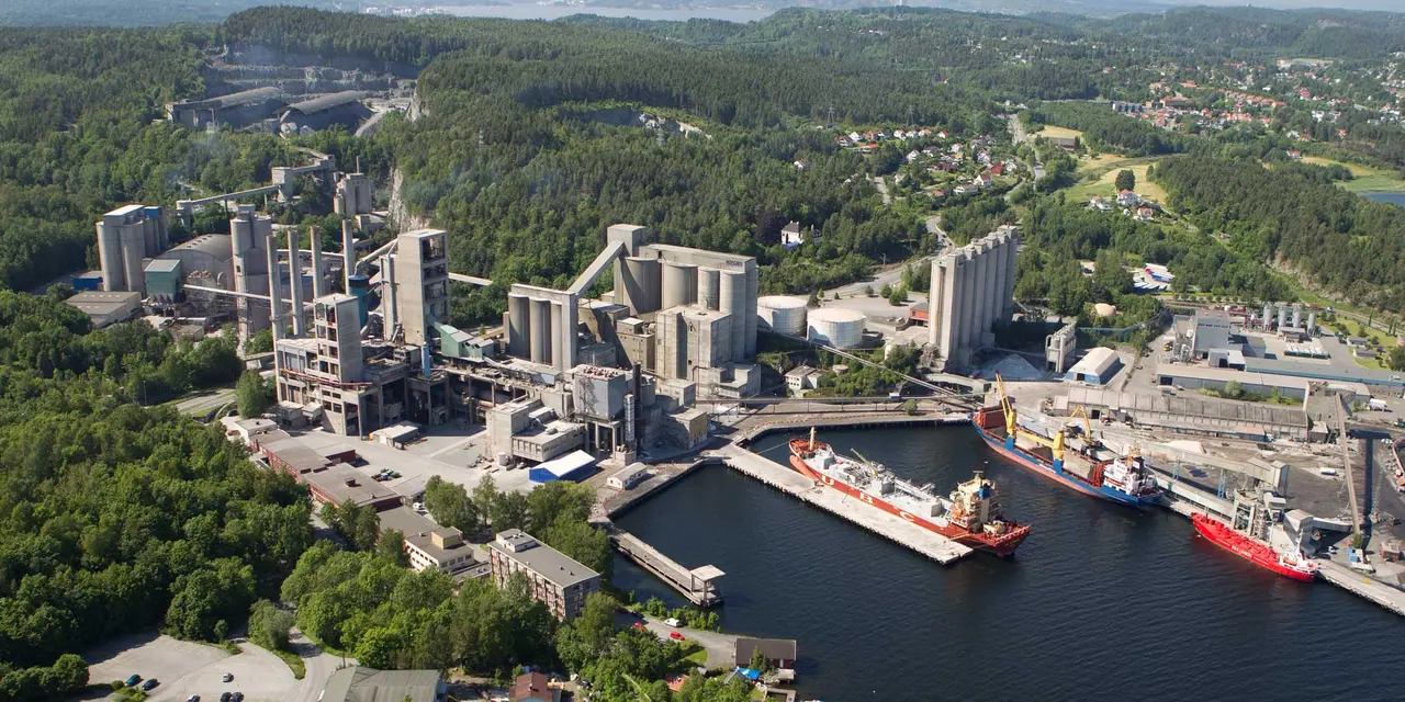 An aerial view of a large industrial facility by the water, showcasing silos, tall buildings, and a docked ship at a pier, surrounded by forest and distant buildings.