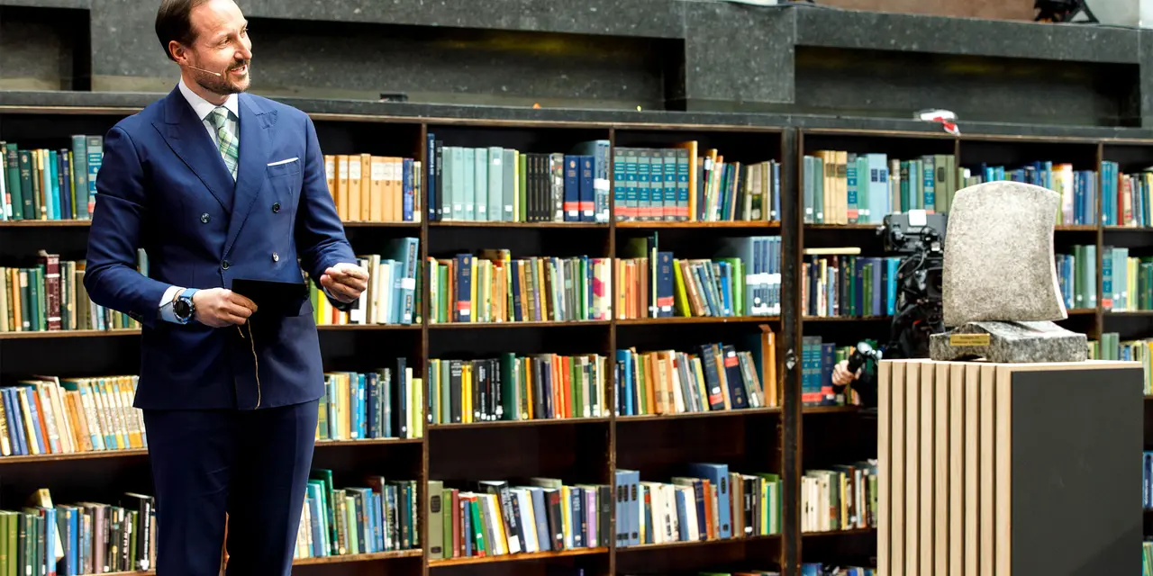 Eine Person in einem blauen Anzug steht vor einem Bücherregal voller bunter Bücher, mit einer abstrakten Skulptur auf einem Podest rechts