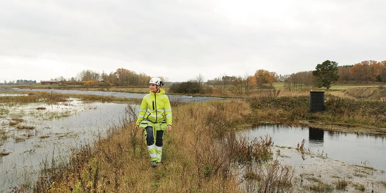 Eine Person in Sicherheitskleidung und mit einem Schutzhelm steht an einem Teich in einem grasbewachsenen Feld, über ihr ist der Himmel bedeckt