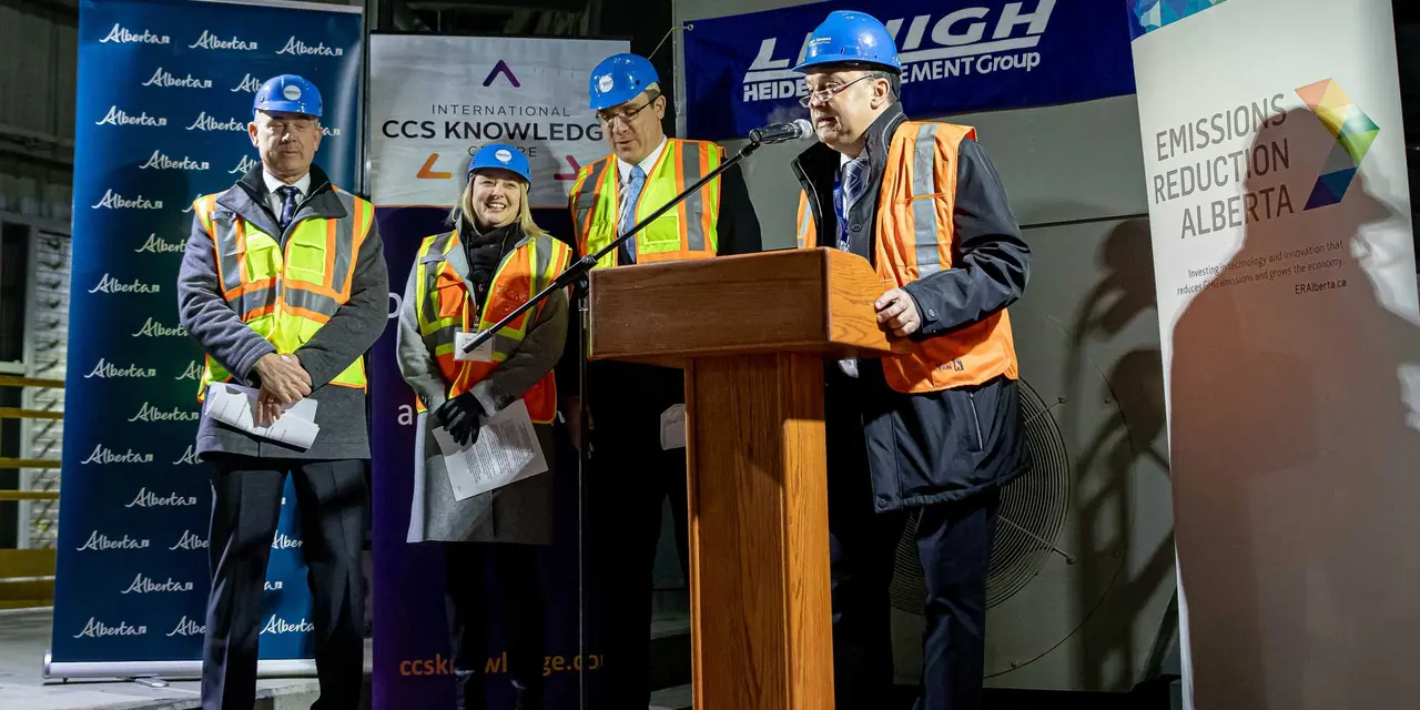 The image shows four individuals standing behind a podium in an industrial environment, their faces not visible. Banners with the logos “CCS KNOWLEDGE,” “HEIDELBERGCEMENT,” and “EMISSION REDUCTION ALBERTA” are displayed in the background.