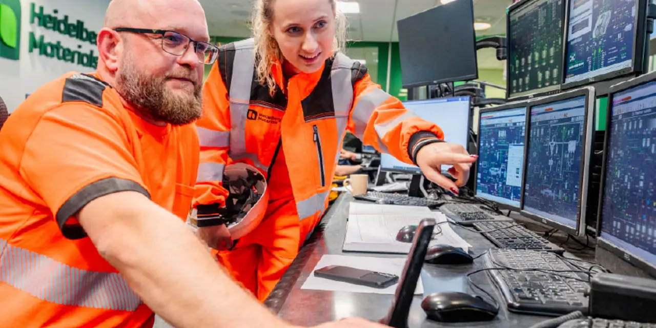 Two employees wearing high-visibility vests are seated at a control desk, monitoring multiple computer screens displaying technical data. The person on the left is holding a mouse, and the person on the right is focused on a screen.