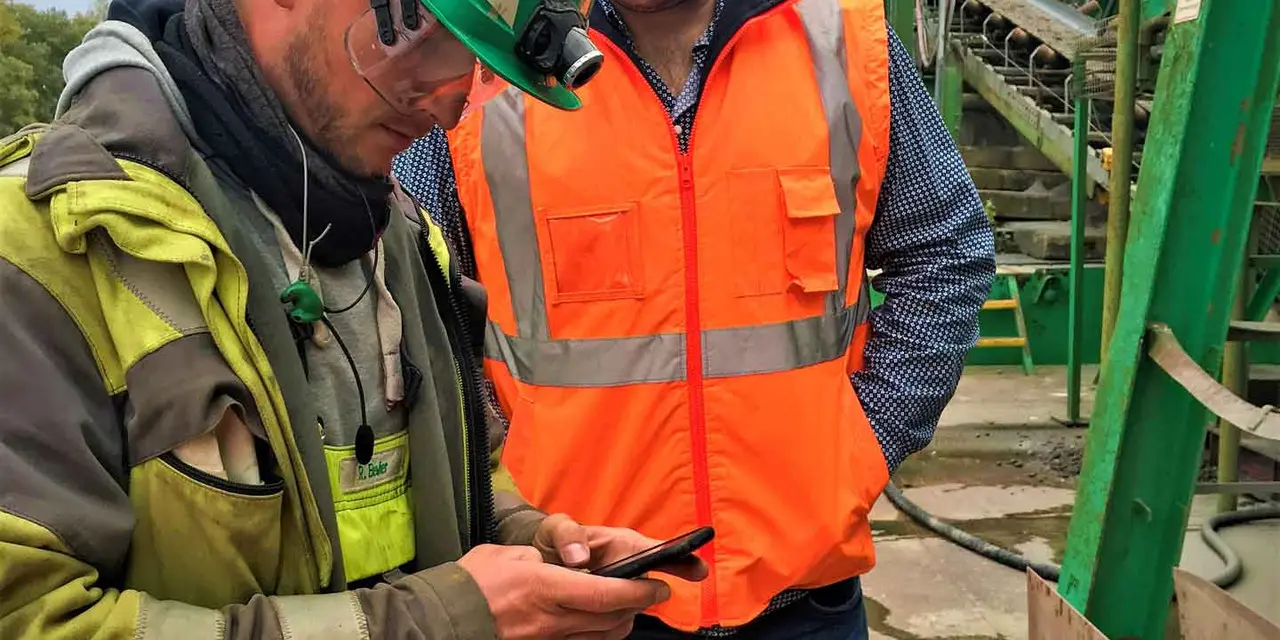 Two workers in safety gear discussing over a walkie-talkie at a construction site with heavy machinery in the background.
