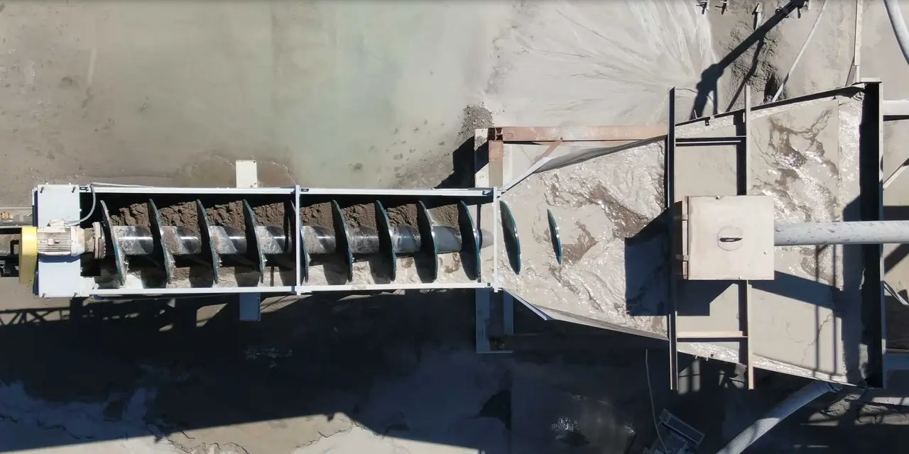 Luftaufnahme von oben eines großen industriellen Spiralförderbandes. Das Band ist mit einer dunklen, schlammigen Substanz gefüllt und entlädt sie auf eine darunterliegende Betonfläche.