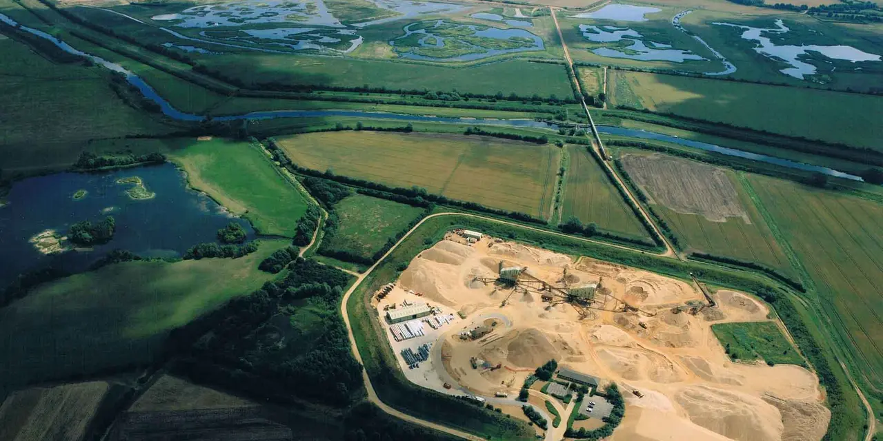 Aerial view of a wetland with a meandering river and a quarry with machinery at work in the lower right, surrounded by lush green fields.