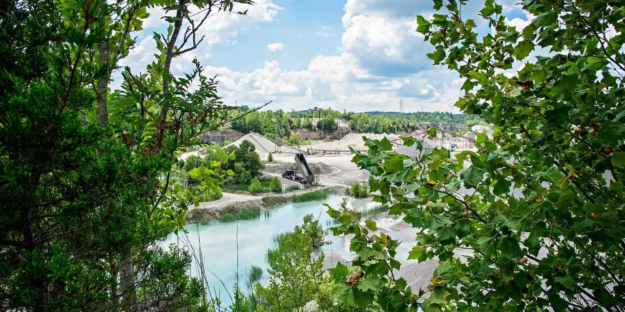 Blick auf einen Steinbruch durch Laub mit einem türkisfarbenen See umgeben von Schotterhaufen und Industrieanlagen.