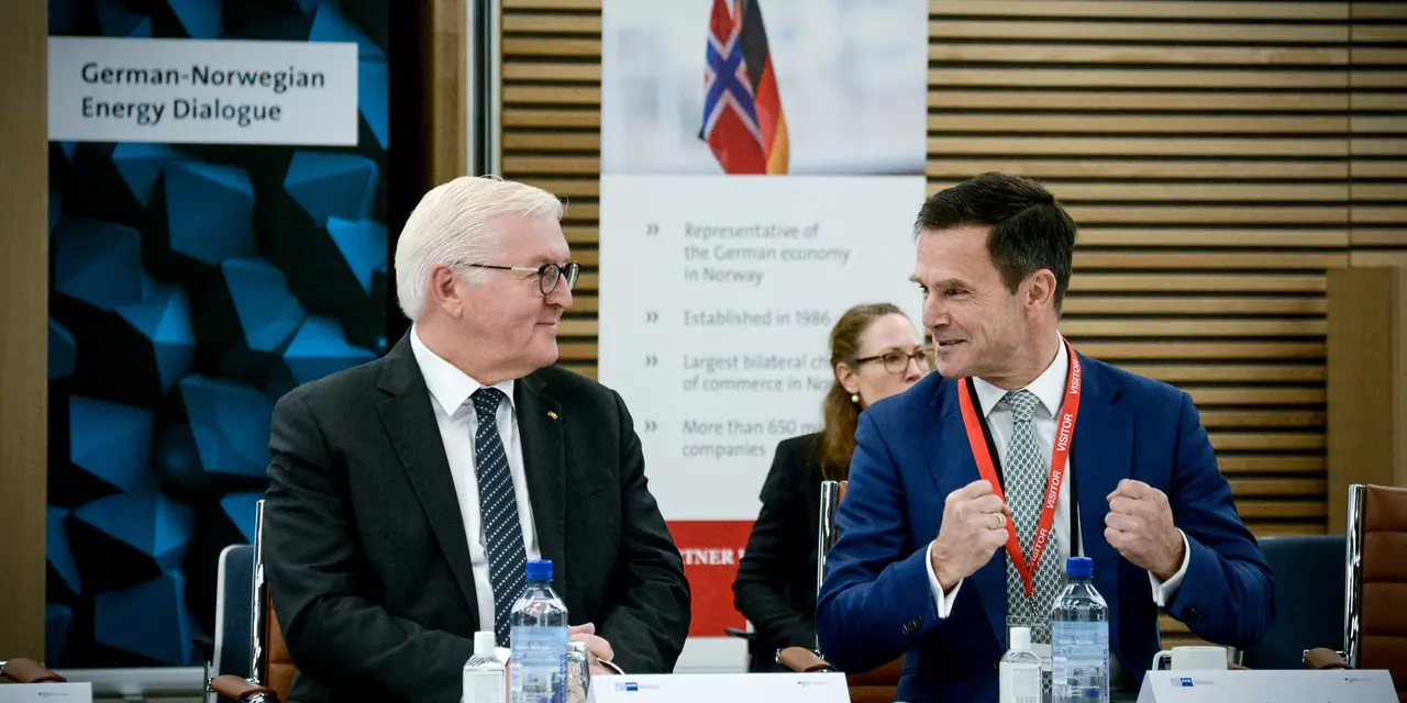 Frank-Walter Steinmeier and Dominik von Achten are seated at a table with microphones, engaged in a formal discussion. A partially visible sign in the background describes the meeting setting.