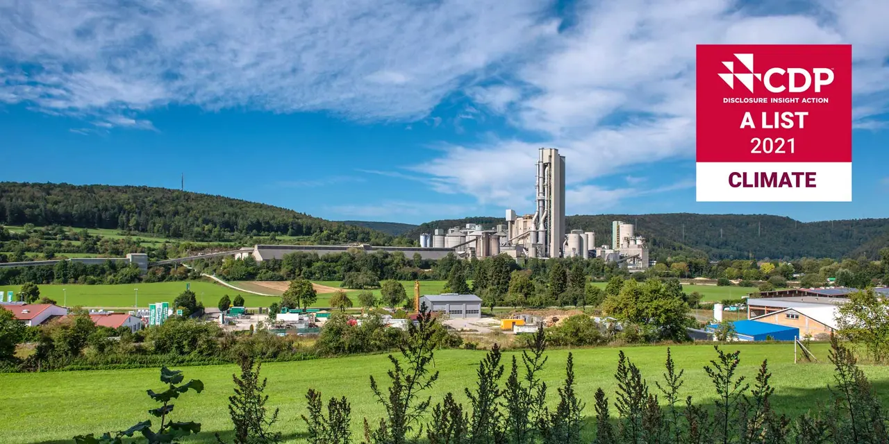 Ein Zementwerk in einer ländlichen Landschaft, umgeben von grünen Feldern und Hügeln. Das Werk hat einen großen, weißen Silo und mehrere andere Strukturen. Das CDP-Logo ist in der unteren rechten Ecke sichtbar, was darauf hinweist, dass das Werk Teil des CDP-Klimawandelprogramms ist.