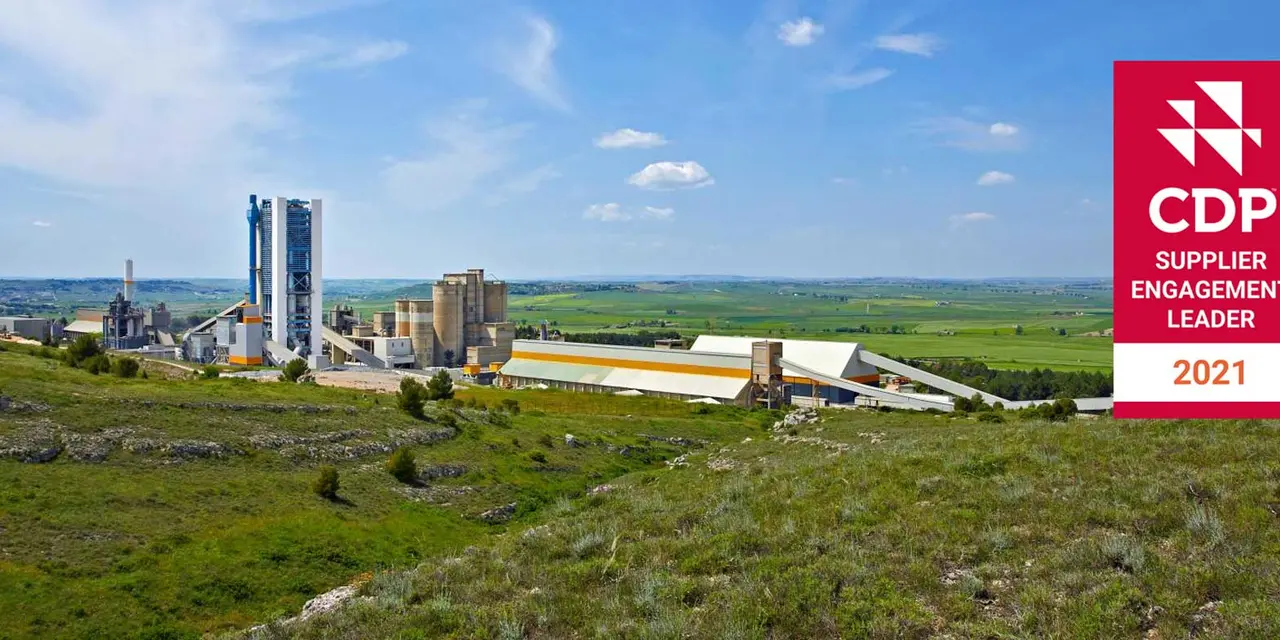 Ein Zementwerk in einer ländlichen Landschaft. Das Werk besteht aus mehreren großen Industriegebäuden und -strukturen, umgeben von grünen Feldern und Hügeln. Ein klarer blauer Himmel mit weißen Wolken ist über dem Werk sichtbar. Im Vordergrund befindet sich ein rotes Banner mit dem CDP-Logo und dem Text "SUPPLIER ENGAGEMENT LEADER 2021".