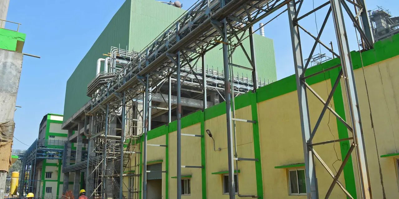 Grün-gelbes Zementwerk mit Metallrohrleitungen rechts und zwei Arbeitern mit Helmen unten unter klarem blauem Himmel.