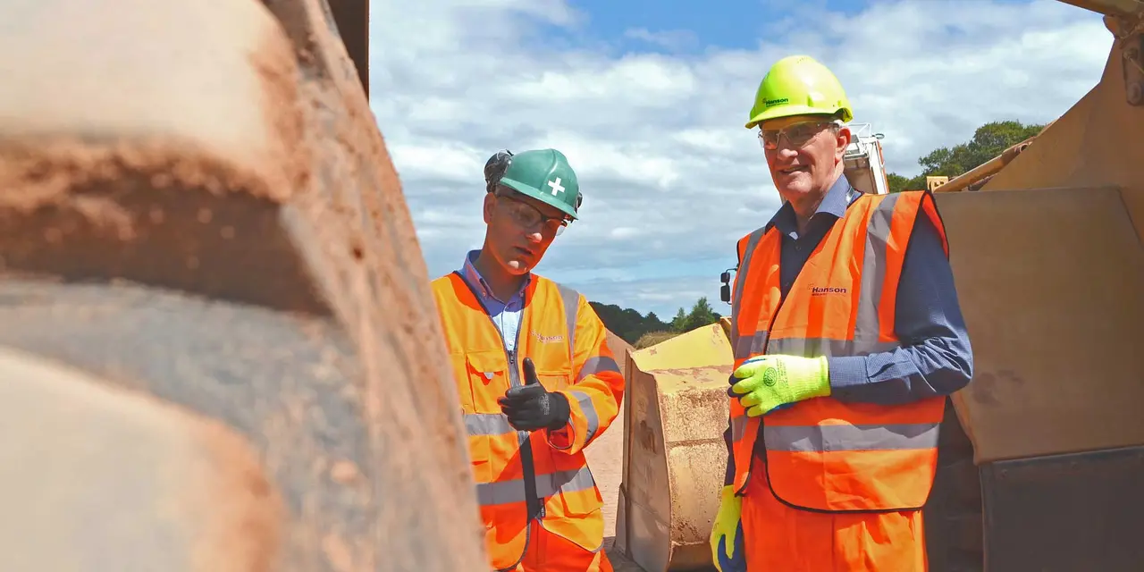 Zwei Personen in orangefarbenen Warnwesten und Schutzhelmen auf einer Baustelle, eine Person hält ein Klemmbrett, beide im Gespräch. Eine unscharfe Mauer ist im Vordergrund zu sehen.