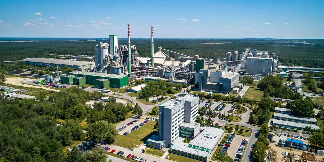 Eine Luftaufnahme eines großen Zementwerks mit mehreren Gebäuden, Silos und Schornsteinen. Das Werk ist von Bäumen und einem Parkplatz umgeben.
