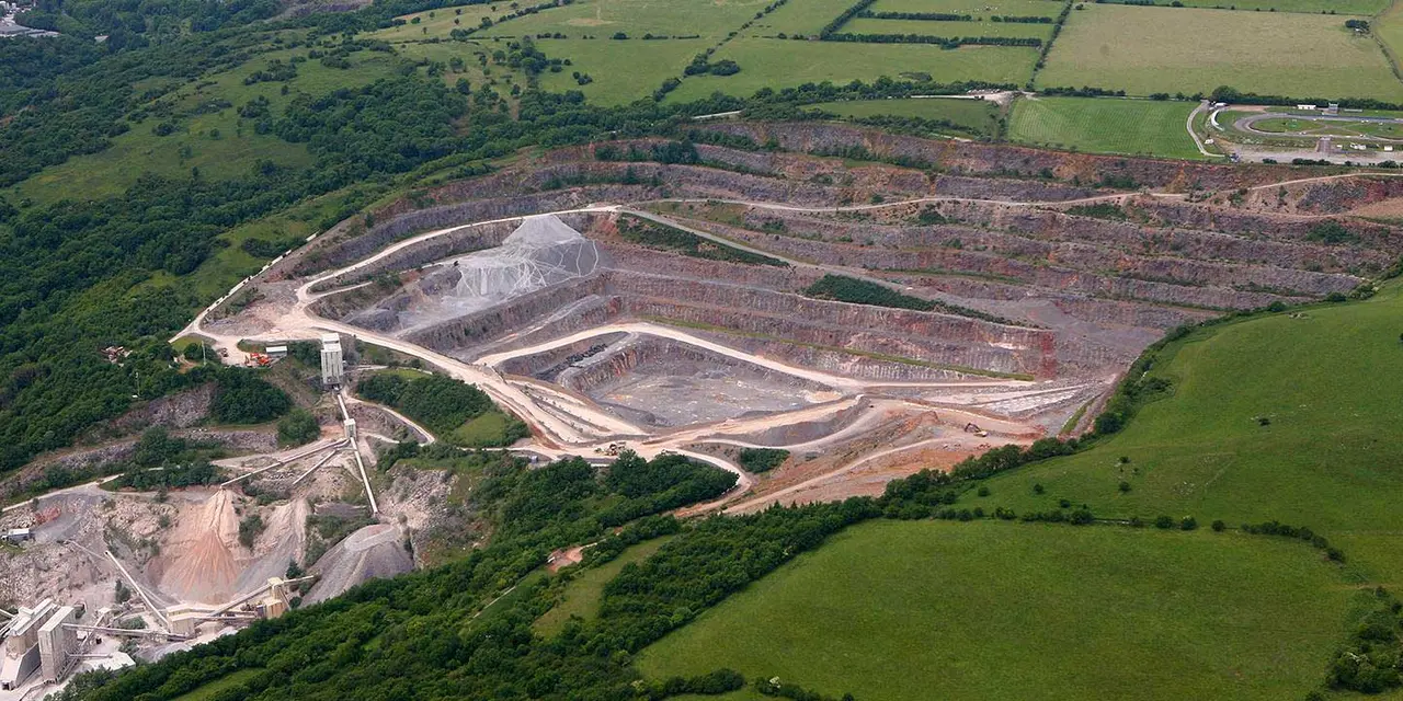 Aerial view of an expansive quarry with terraced excavation levels, surrounded by lush greenery and connected by a winding road.