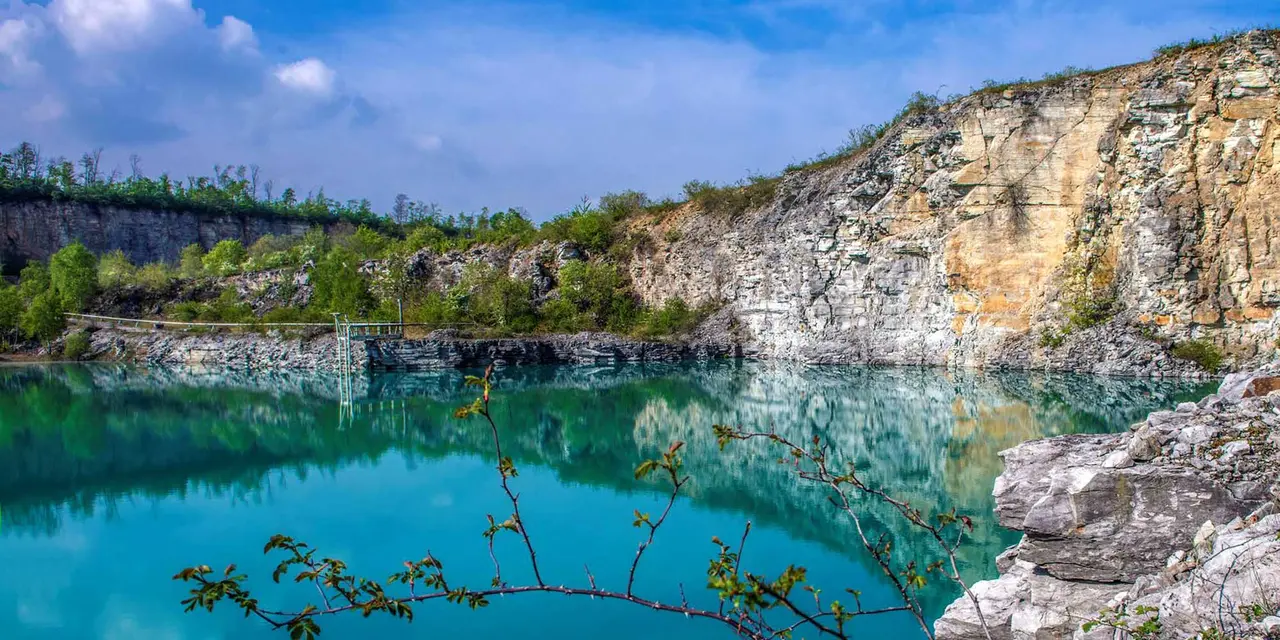 Scenic view of a clear blue lake surrounded by rocky cliffs with lush green vegetation under a partly cloudy sky.