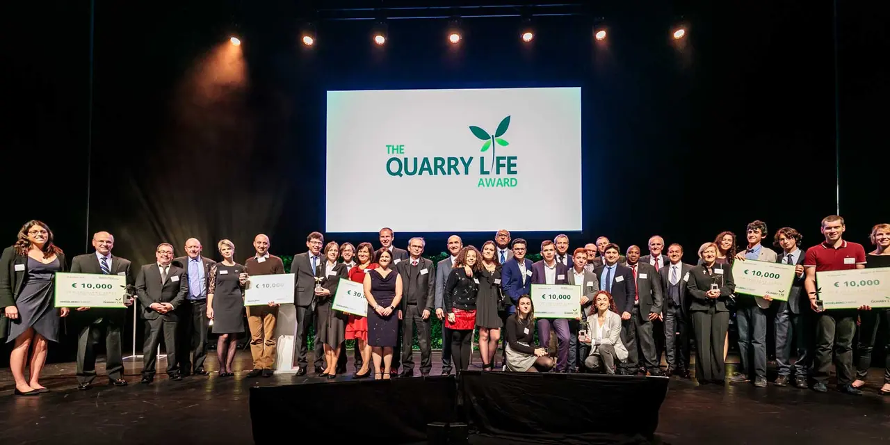 Award recipients lined up on stage at ‘The Quarry Life Award’ ceremony holding checks with prize amounts, with event logo displayed on screen behind.