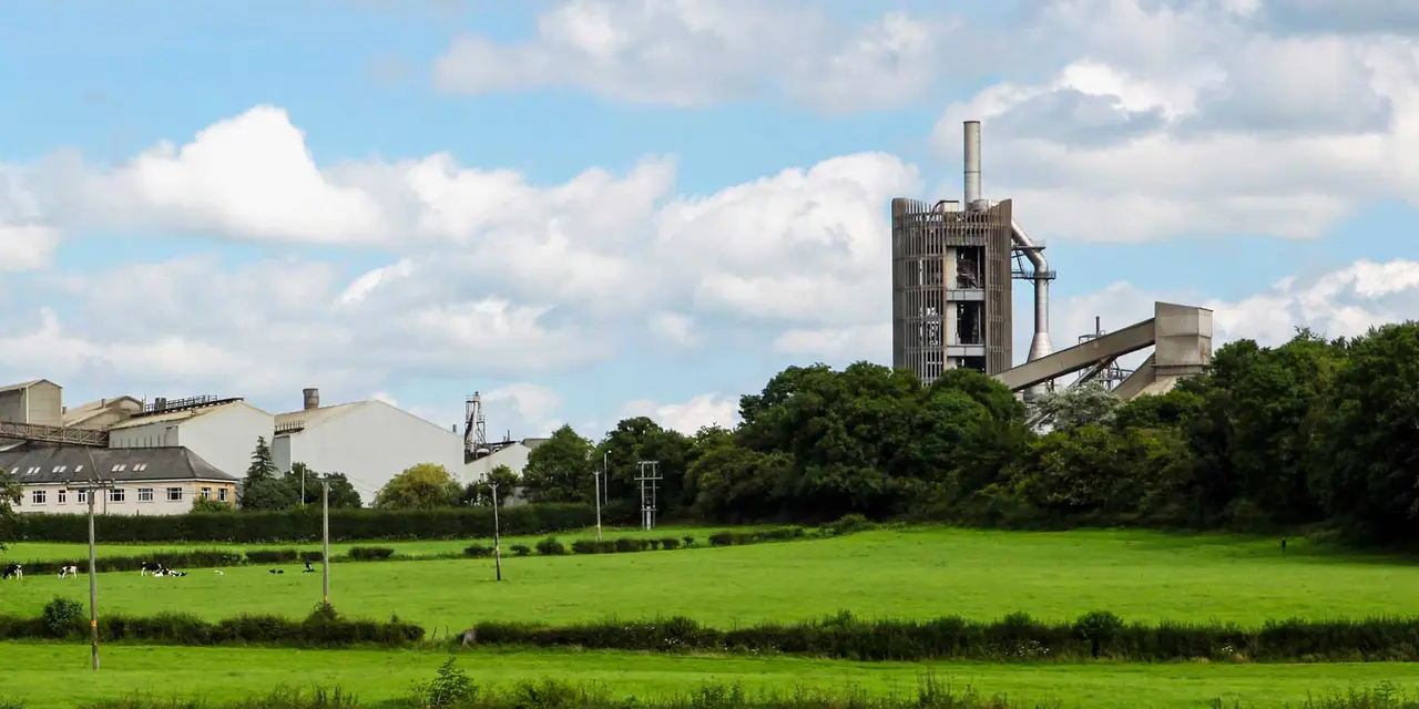 Ein Zementwerk mit einem großen Schornstein und mehreren Gebäuden neben einem grünen Feld unter einem teilweise bewölkten Himmel.