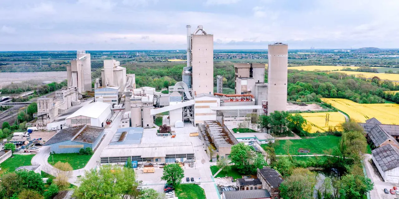 An aerial view of a large cement plant with multiple buildings and silos, surrounded by green fields and a nearby town.