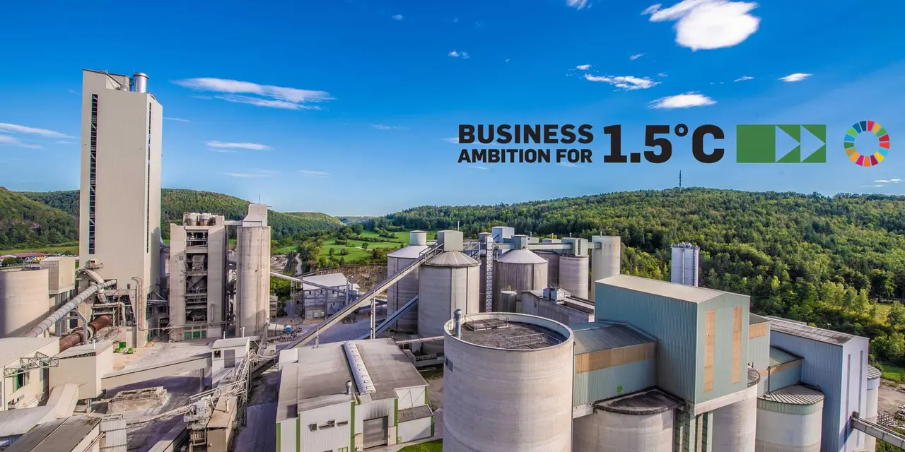 A cement plant with multiple silos and buildings, surrounded by green hills and a blue sky. The words "BUSINESS AMBITION FOR 1.5°C" and the United Nations logo are visible in the top right corner.