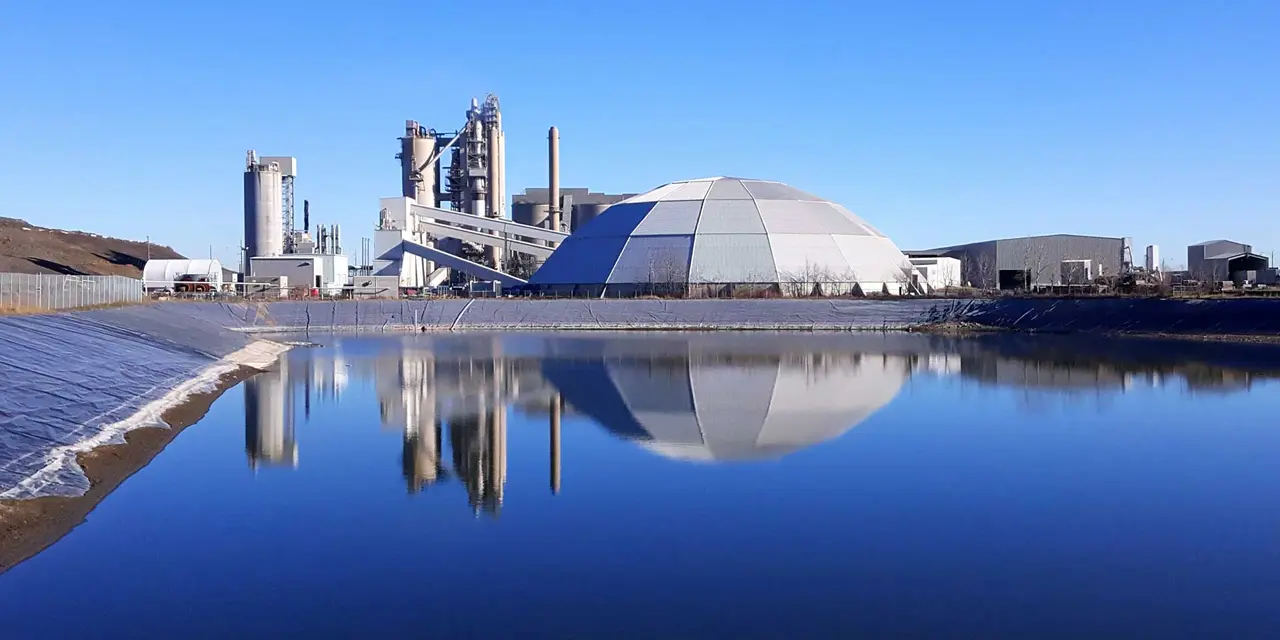 A large cement factory with tall chimneys and a large, dome-shaped building reflected in a body of water. A smaller building is visible to the left.