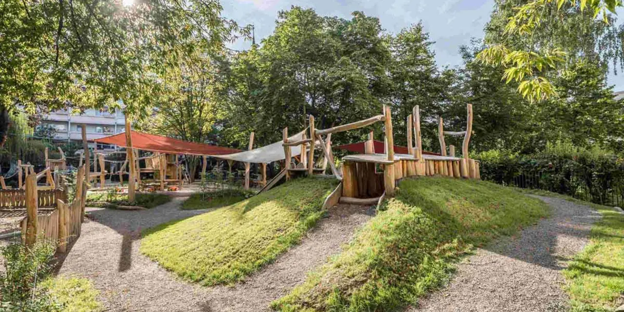 Playground featuring wooden structures, grassy mounds, and gravel pathways, surrounded by trees and greenery.