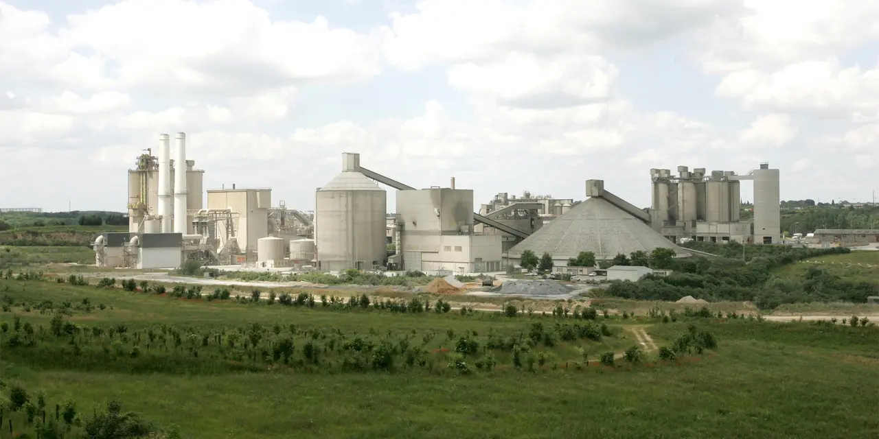 Industrielle Zementanlage mit Silos inmitten grüner Felder unter bewölktem Himmel