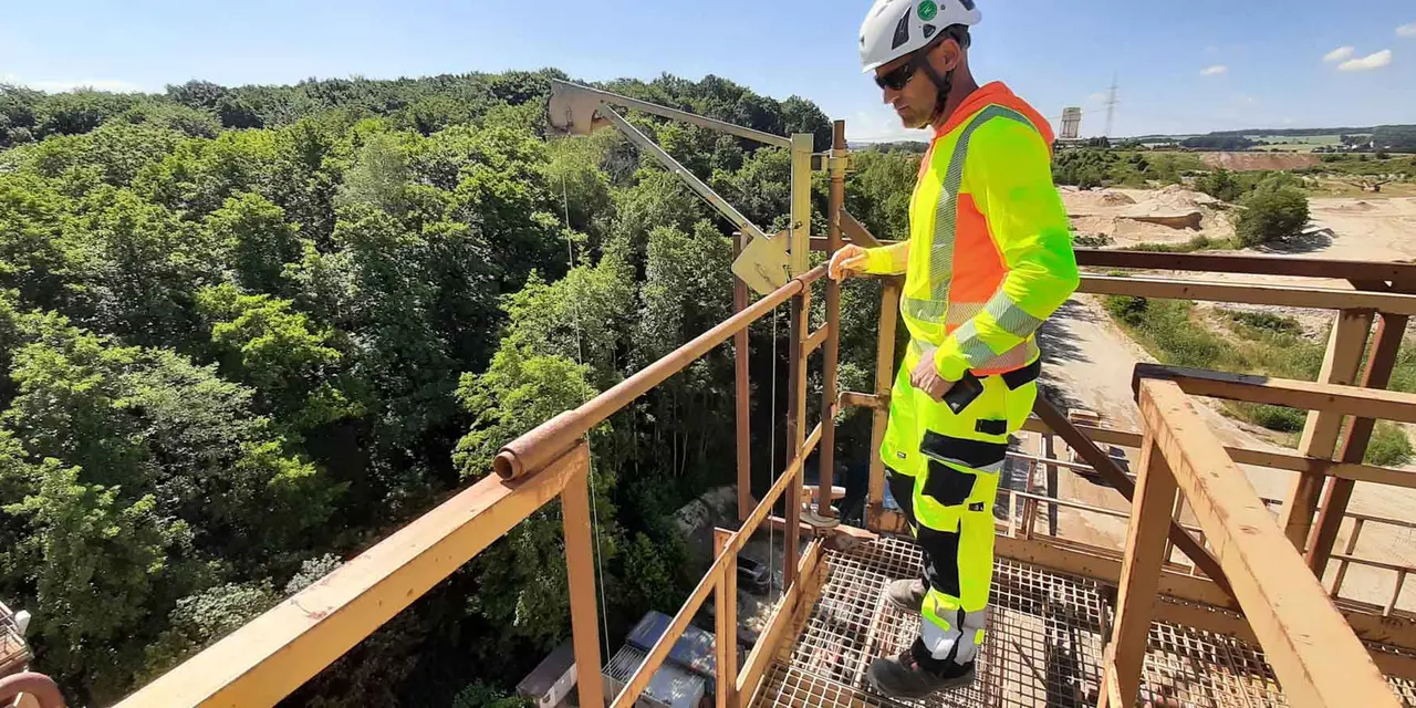Man in protective clothing stands on a scaffolding, in the background forest and the area of a gravel plant