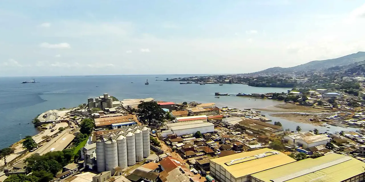 An aerial view of a coastal city with a port and industrial area. There are several buildings, including a silo, and a large body of water with ships in the background.