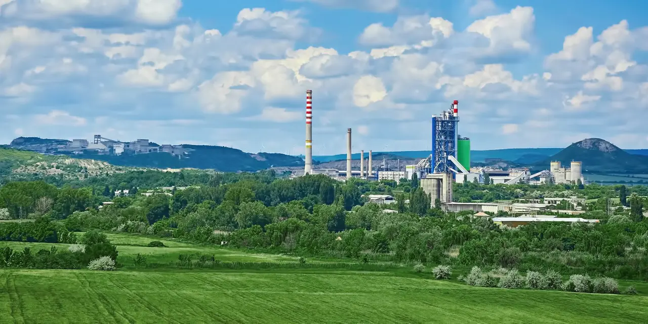 A light green field in front of a cement works, cloudy sky above