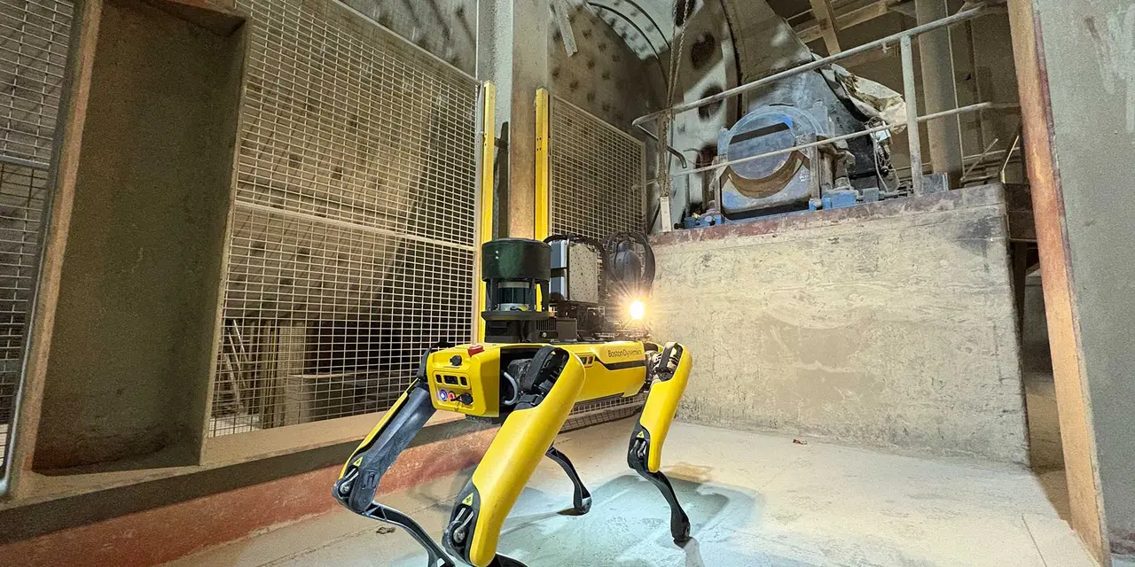 A yellow robotic dog stands in a cement plant with machinery in the background.