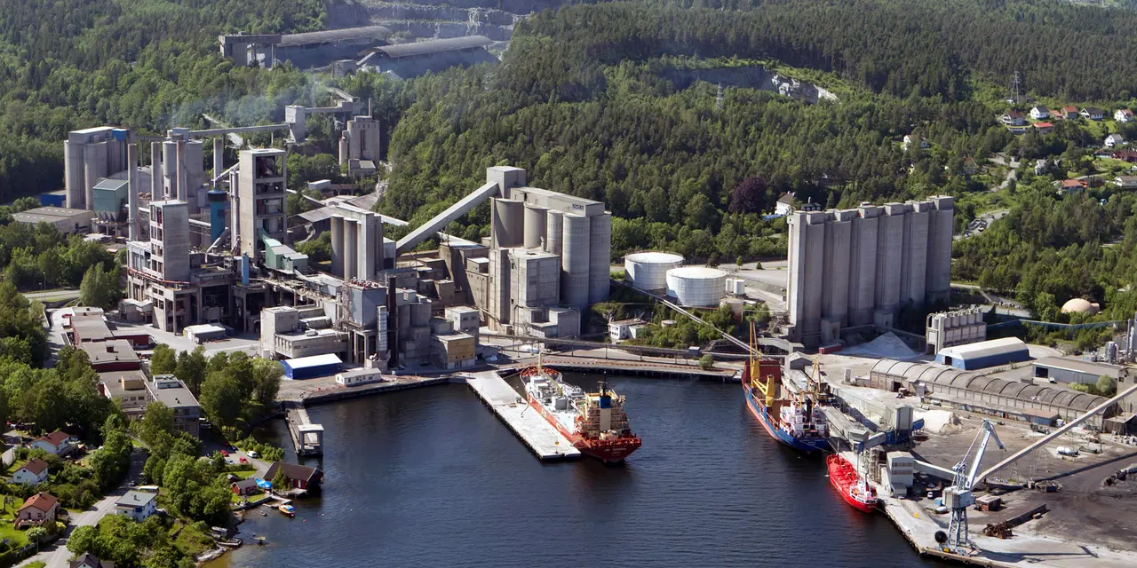 Aerial view of a industrial port with a cement plant, silos, towers, and piping on the waterfront, surrounded by forested hills.