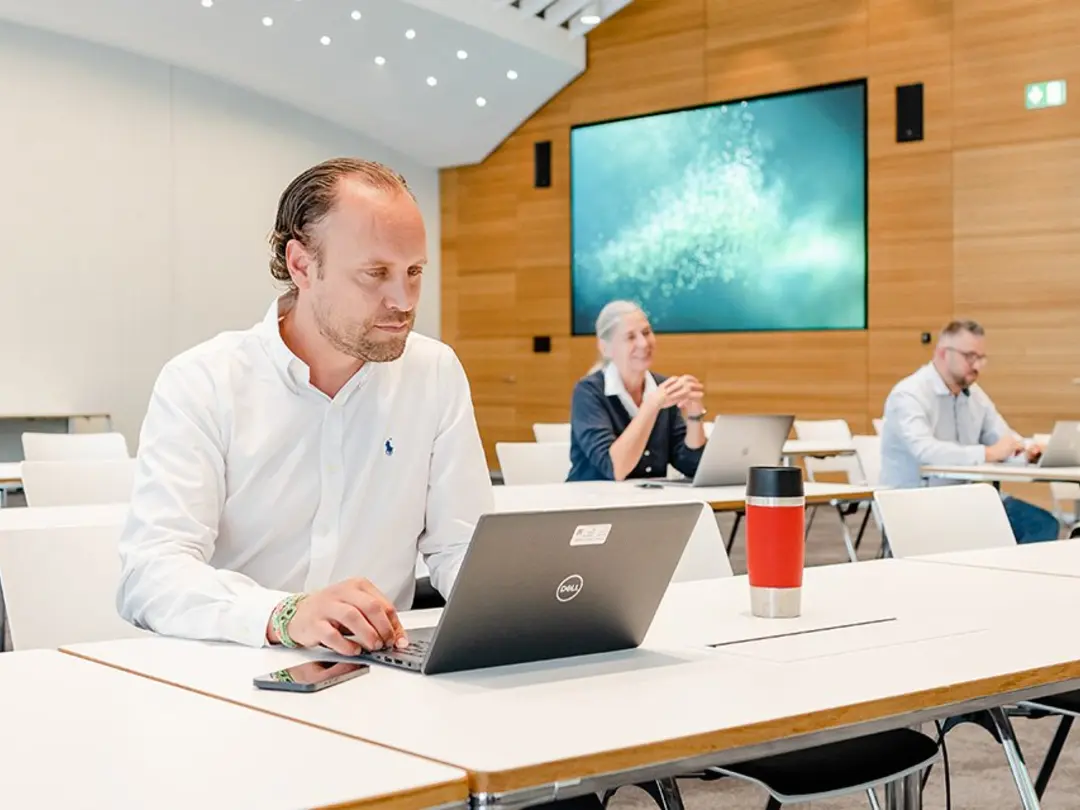 Ein heller Seminarraum mit weißen Tischen und Stühlen. Im Vordergrund sitzt ein Mann in einem weißen Hemd, der auf einem Laptop arbeitet. Im Hintergrund sind zwei weitere Personen an separaten Tischen, eine davon mit einem Smartphone. An der holzverkleideten Rückwand hängt ein großer Bildschirm, der ein Bild eines blauen Ozeans zeigt.