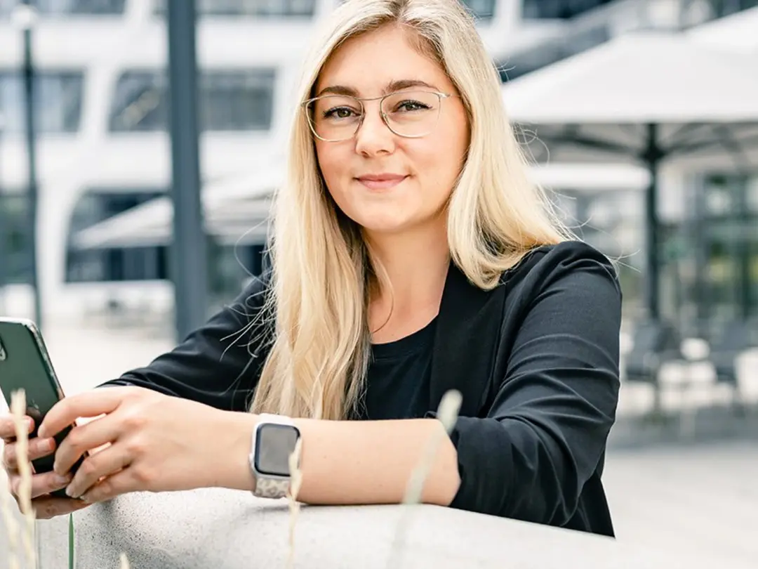 A woman is holding a mobile phone, looking and smiling into the camera
