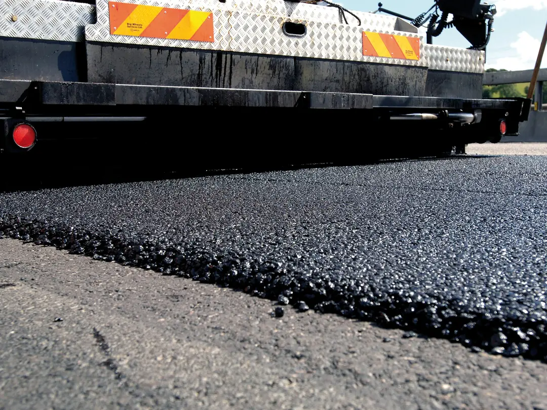 Ein Baufahrzeug trägt dunkelgraue Masse auf eine Straße auf.