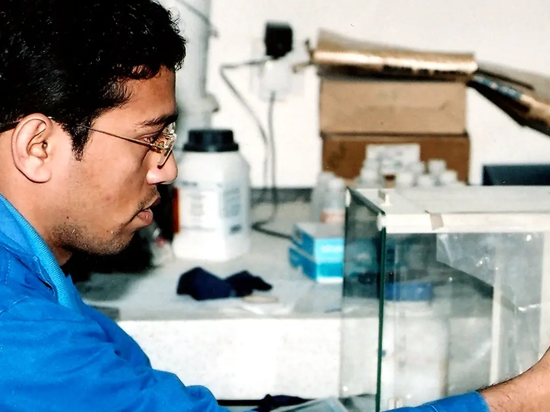 Person im blauen Laborkittel bei der Arbeit mit Geräten an einer Holztafel in einer Laborumgebung, umgeben von Glasbehältern, Kartons und anderen Laborgeräten.