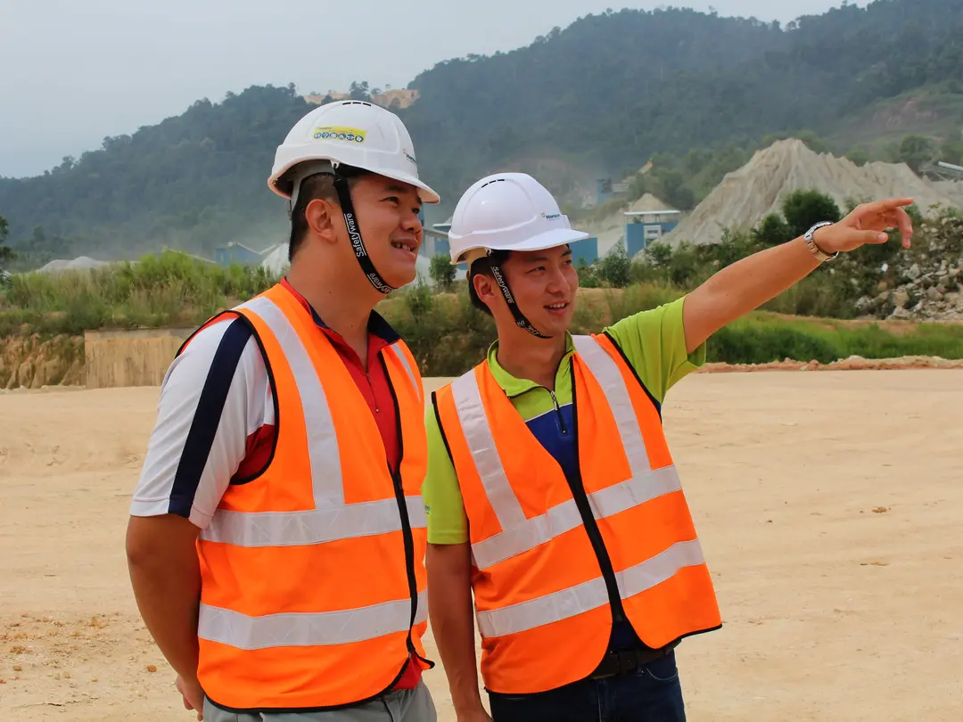 Zwei Personen mit Warnwesten und Schutzhelm auf einem Steinbruch, eine zeigt in die Ferne