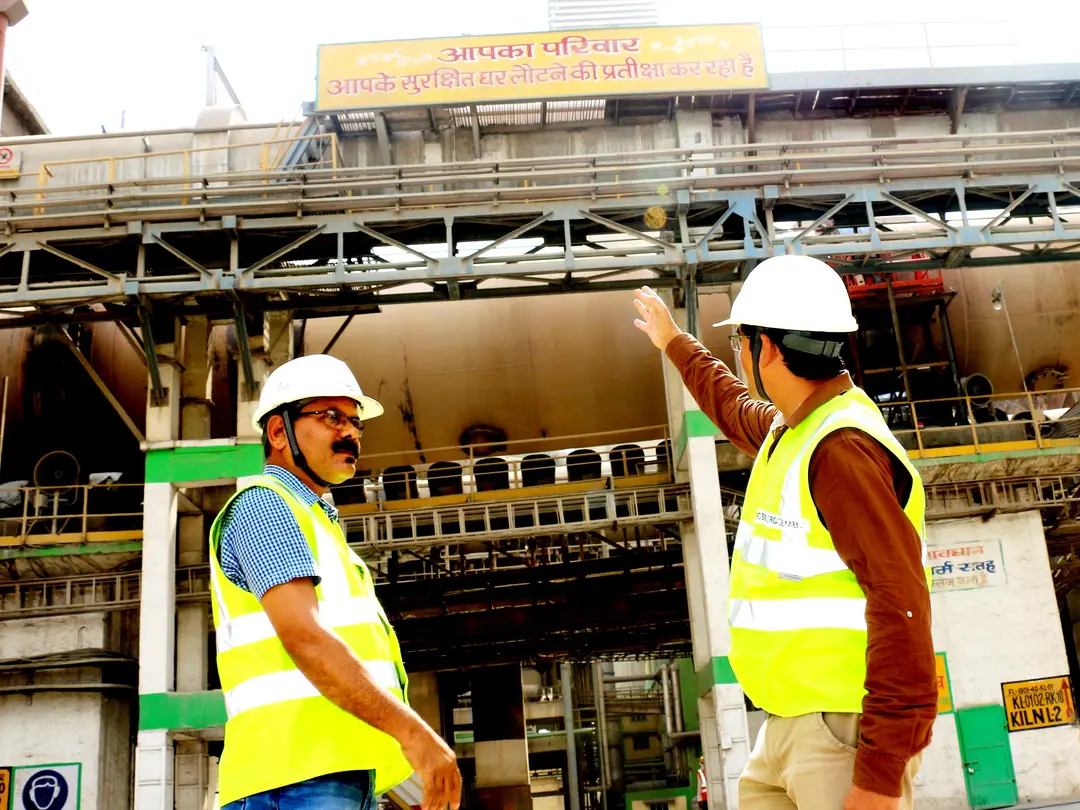 Zwei Bauarbeiter in Sicherheitswesten und Helmen stehen vor einer Industrieanlage, einer zeigt gestikulierend auf die Struktur.