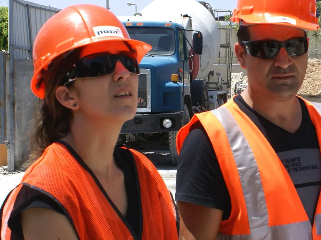 Personen in Sicherheitskleidung und Helmen bei einer Baustelle mit Lkws im Hintergrund, möglicherweise im Zusammenhang mit Zement- oder Betonproduktion.