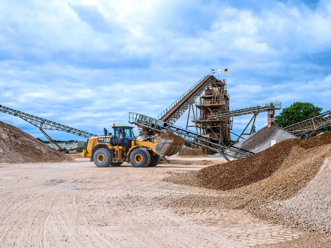 Bagger in einem Kieswerk lädt Sand auf; im Hintergrund Förderbänder, eine Kiesaufbereitungsanlage und Sandhügel; blauer Himmel mit Wolken.