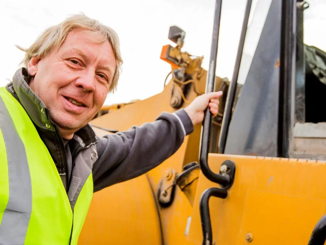 Mann in Warnweste zeigt auf die Tür eines gelben Baufahrzeugs; im Hintergrund ist ein Lenkrad sichtbar.