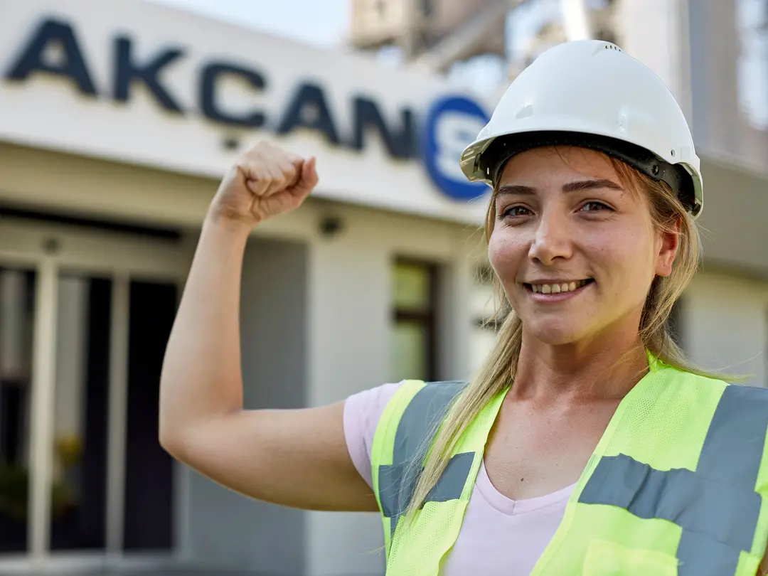 Frau in Sicherheitsweste und Schutzhelm zeigt eine starke Geste mit erhobenem Arm im Vordergrund, Gebäude mit dem Schriftzug „AKCAN“ im Hintergrund.